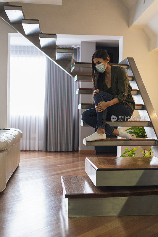 一名妇女坐在家里的楼梯上，态度非常无聊，因为她是COVID - 19阳性，必须用口罩隔离，以避免病毒传播。